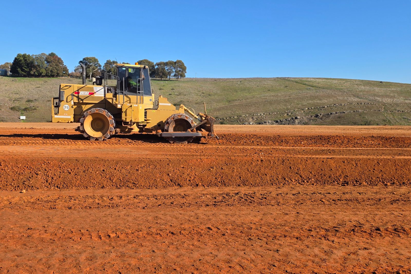 A yellow padfoot compactor rolling the red earth subgrade of a new road corridor in front of a green hill.