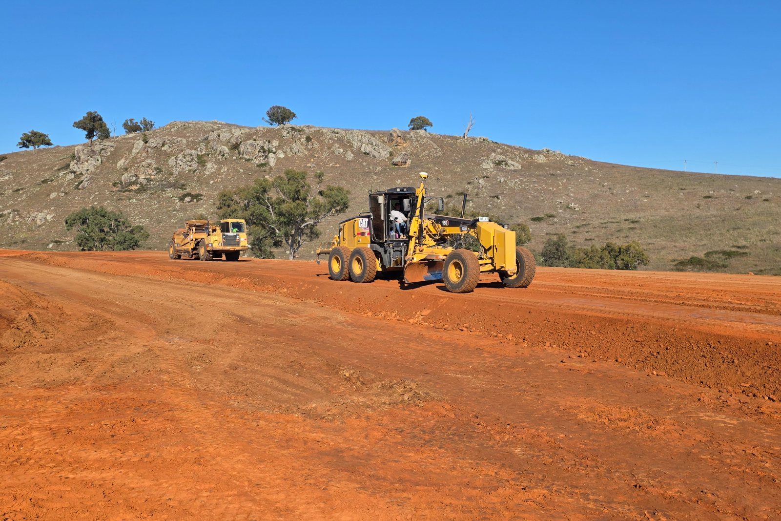 A grader and a scraper working a wide bank of red-brown earth for a new road, with a rocky grass hill behind.