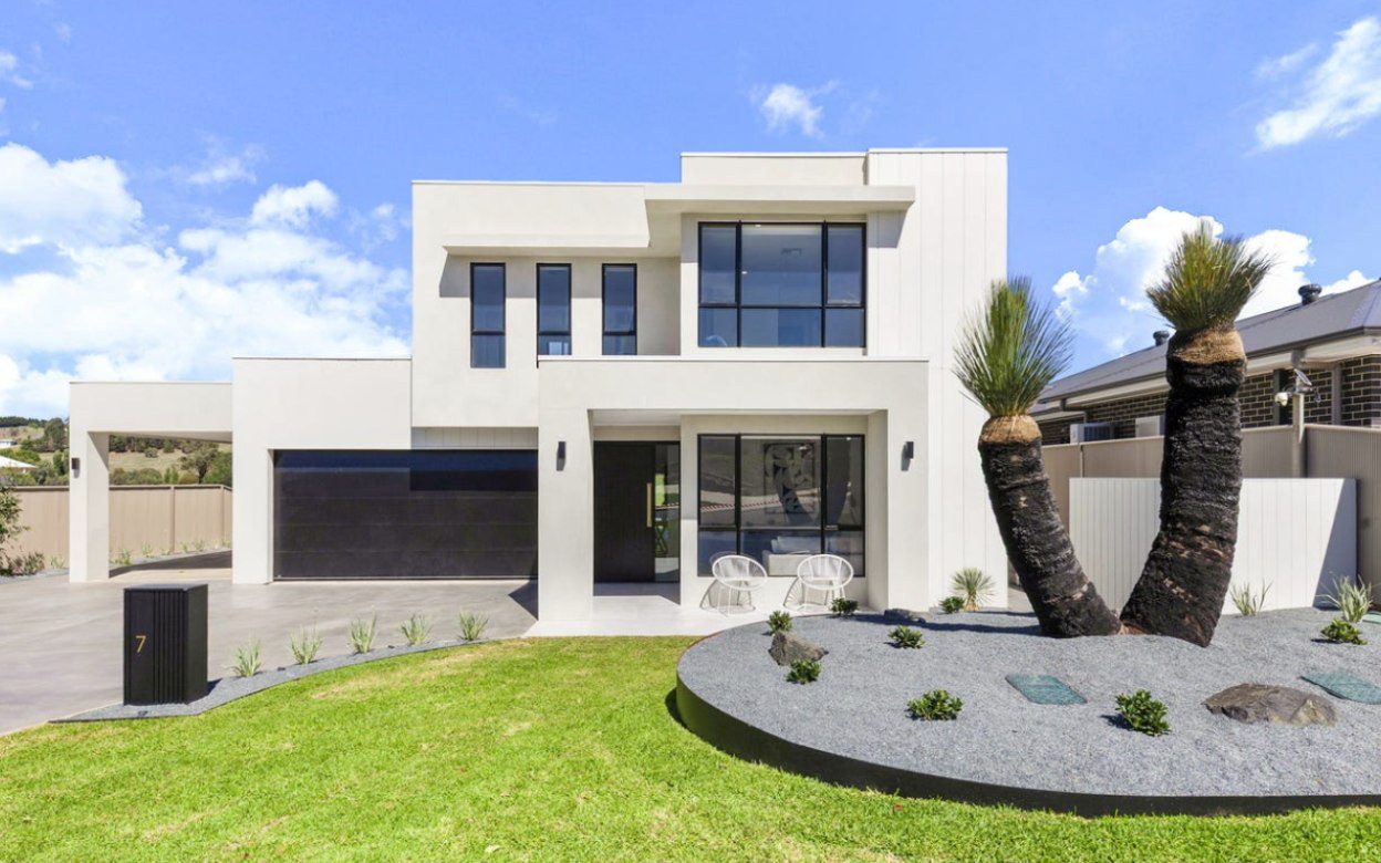 A completed two-storey home at Yarrah in white render with black-framed windows, a double garage, lawn and a gravel garden bed planted with grass trees.