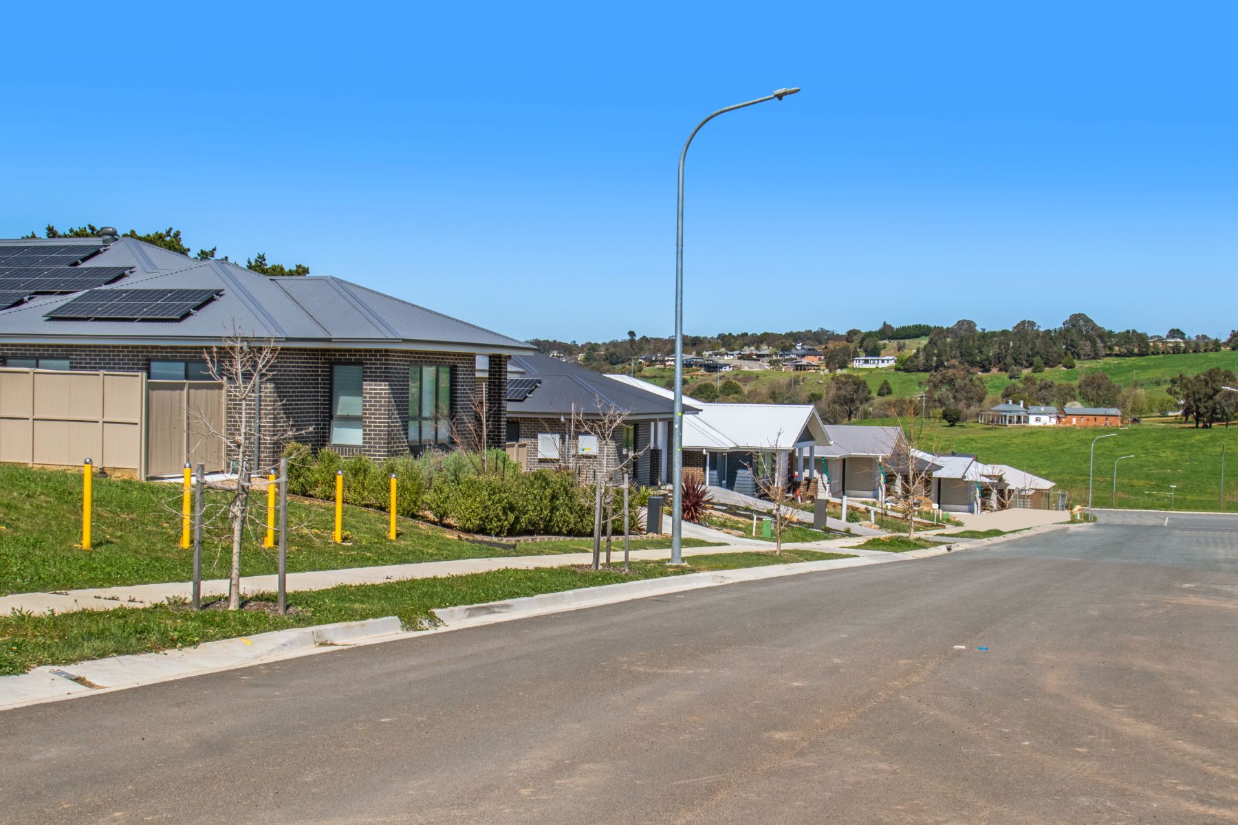 A sealed street lined with new brick and weatherboard homes, young street trees, a footpath and a street light under a clear blue sky.