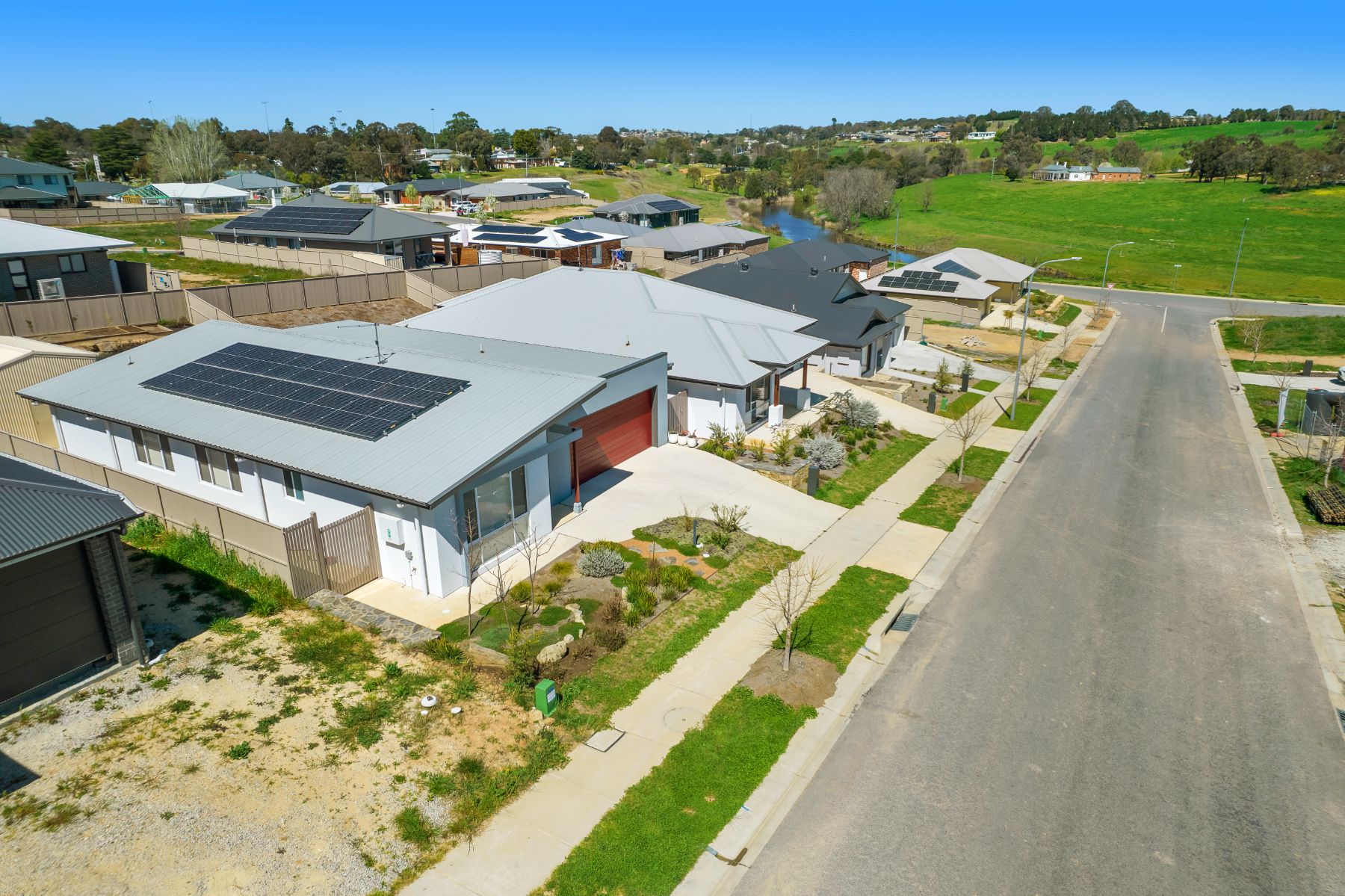 Aerial view of a street of completed single-storey homes with solar panels, landscaped front gardens and a footpath, with a river and green paddocks in the distance.