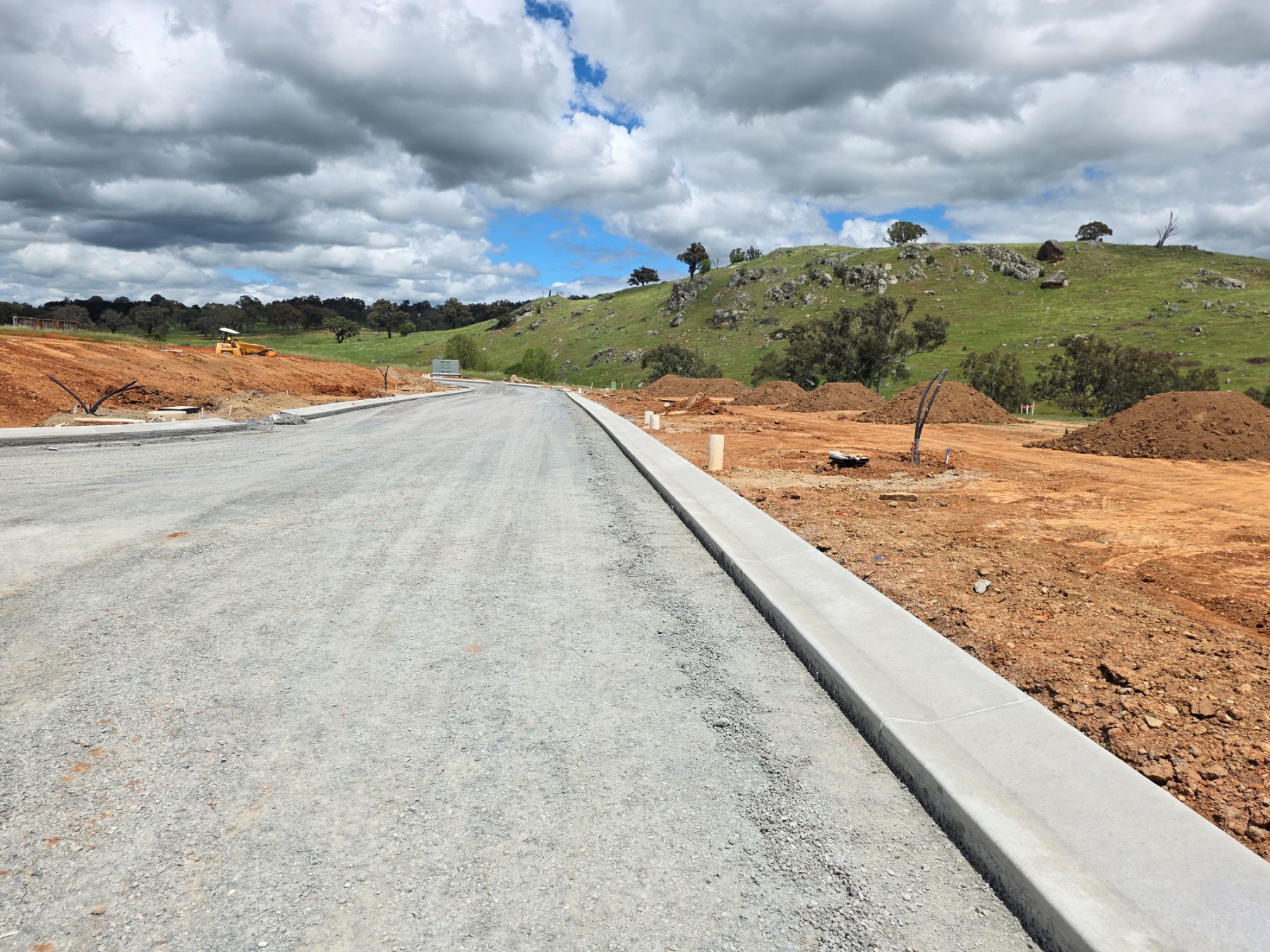 A new road with a compacted gravel subbase and a freshly poured concrete kerb, bare red earth beside it and a rocky green hill behind.