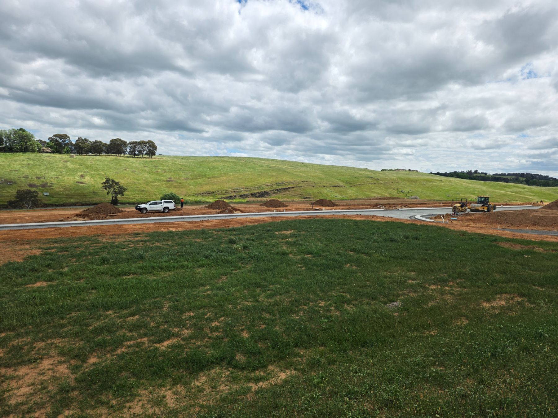 A grassed area in the foreground, a new road with kerbs beyond it, a utility vehicle and earth mounds, and a roller working near an intersection.