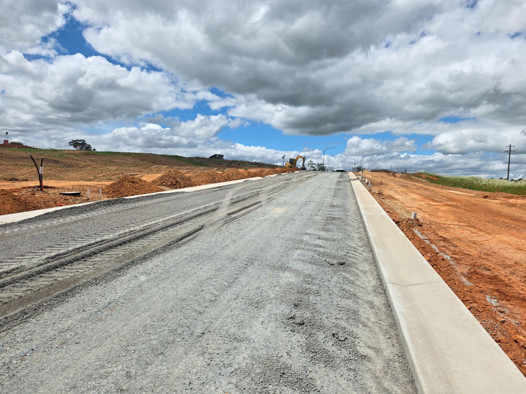 A wide gravel road rising towards an excavator, with a concrete kerb on the right and red earth mounds on the left under heavy cloud.