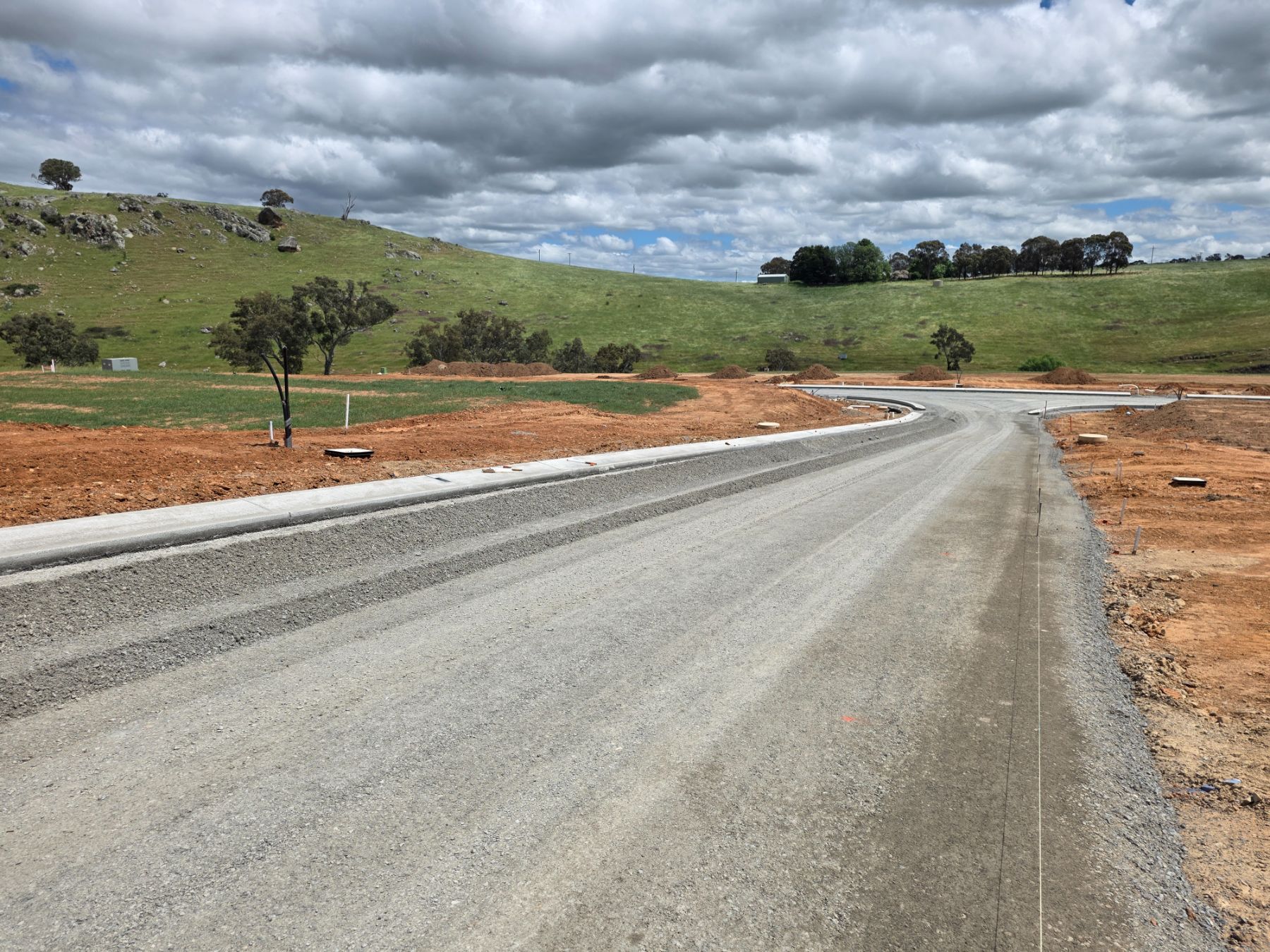 A new gravel-surfaced street with a concrete kerb on one side, curving to a T-intersection, with green hills and cloudy sky behind.