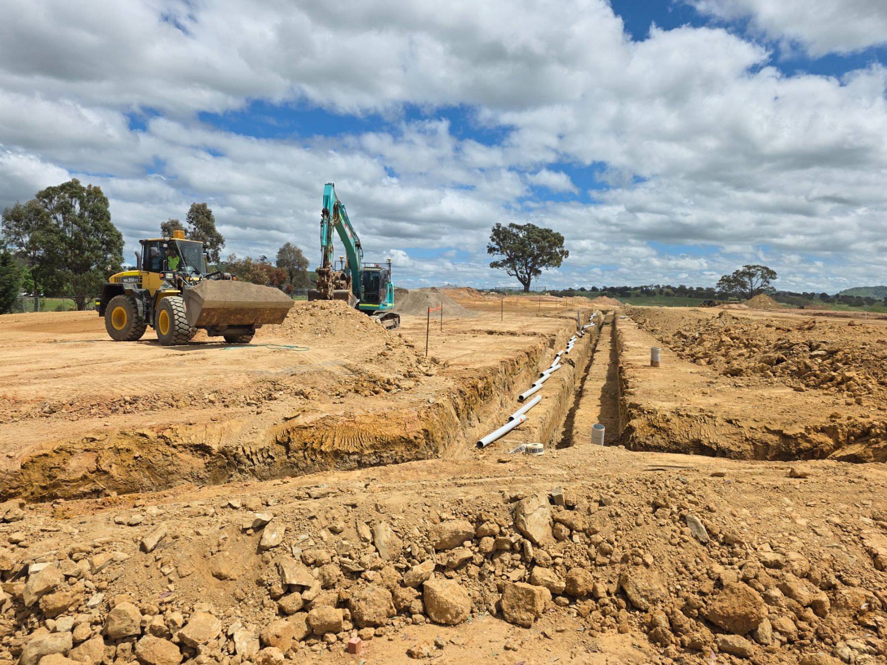 A wheel loader with a full bucket and an excavator beside a long open trench, with white pipe sections laid along its edge.