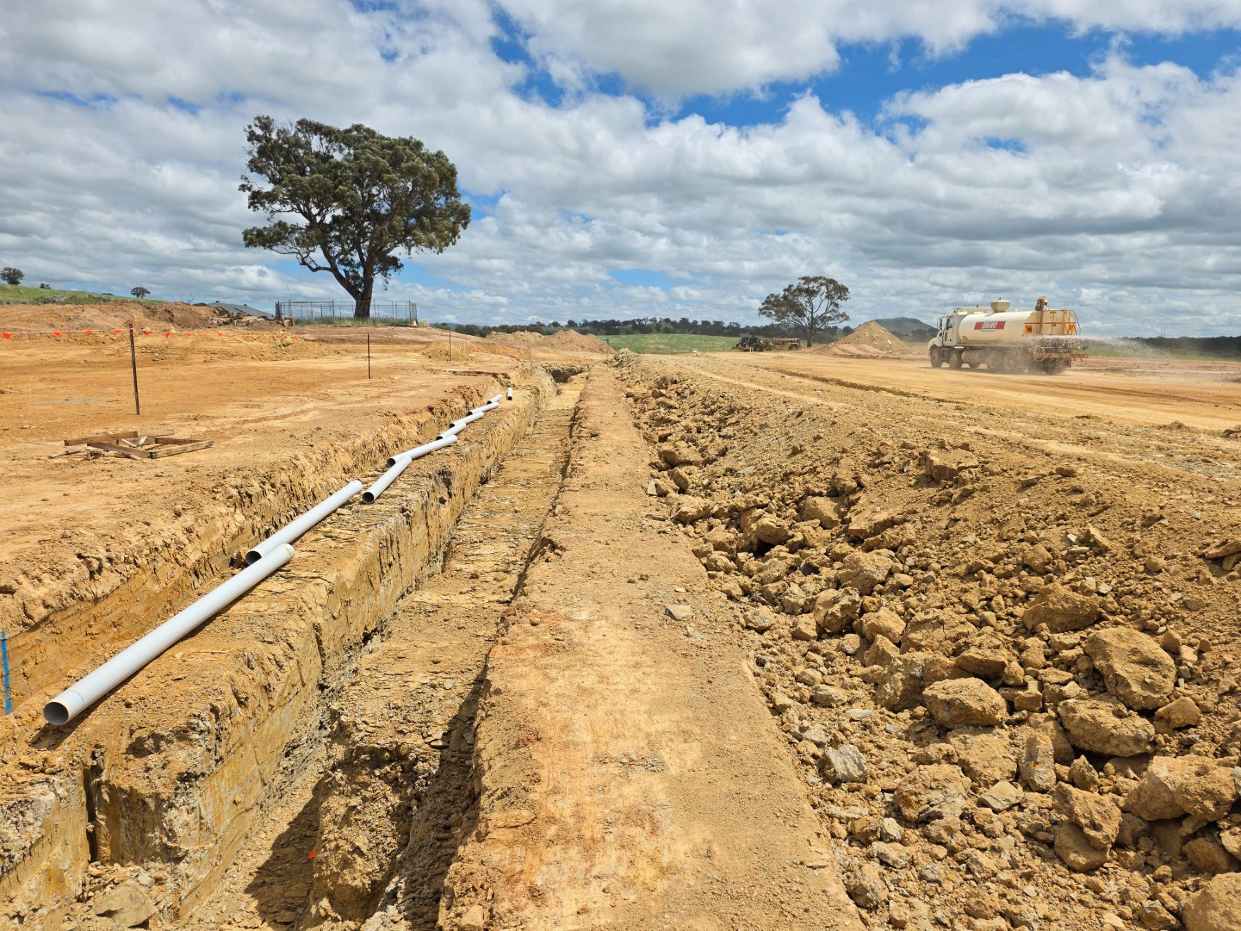 White pipe sections lying along the edge of a trench in dry orange ground, with a water truck spraying the haul road and a lone tree behind.
