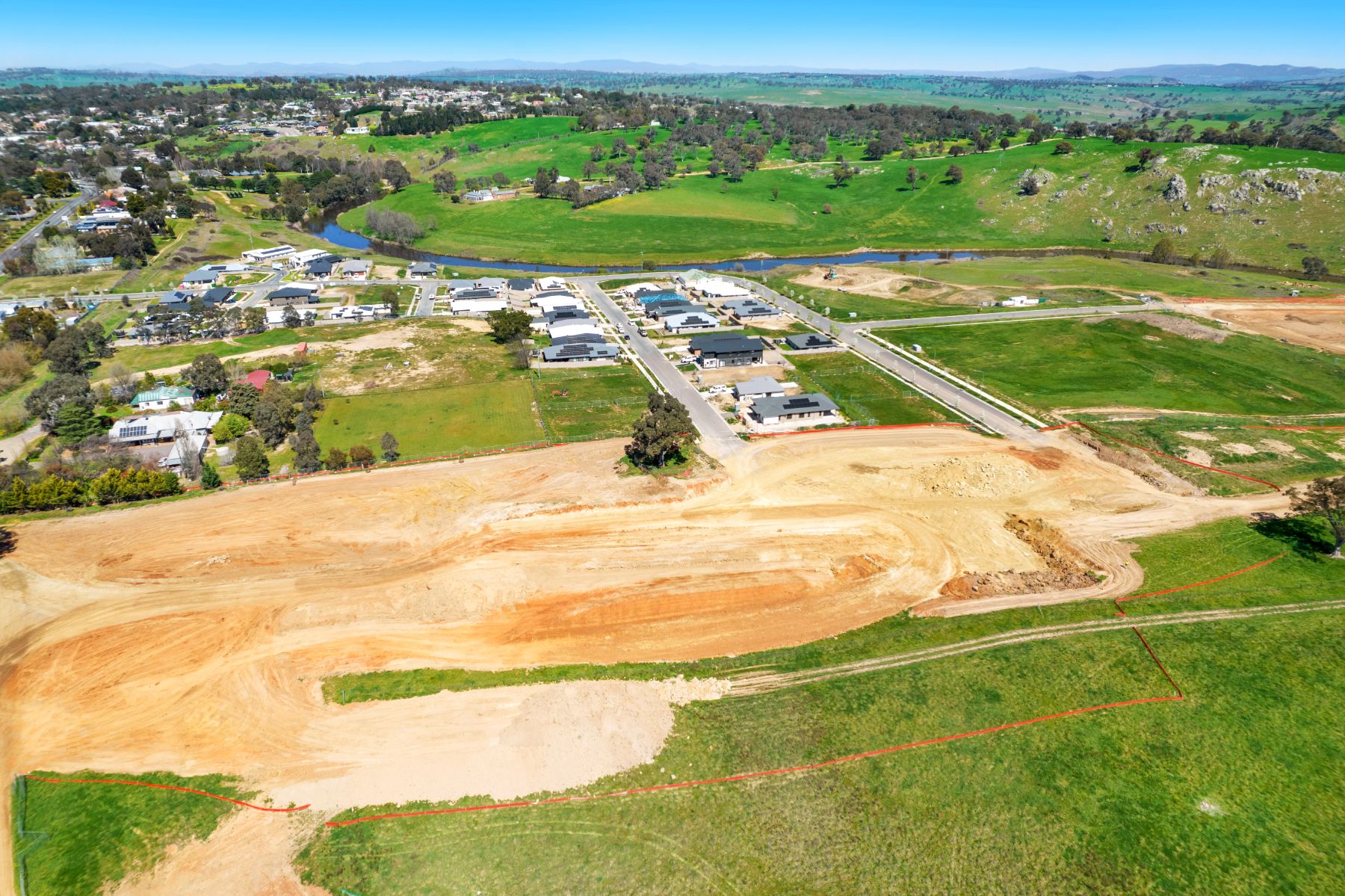 Aerial view of a large area of cleared, shaped earth bordered by orange safety fencing, next to a street of new homes, with a river and green hills beyond.