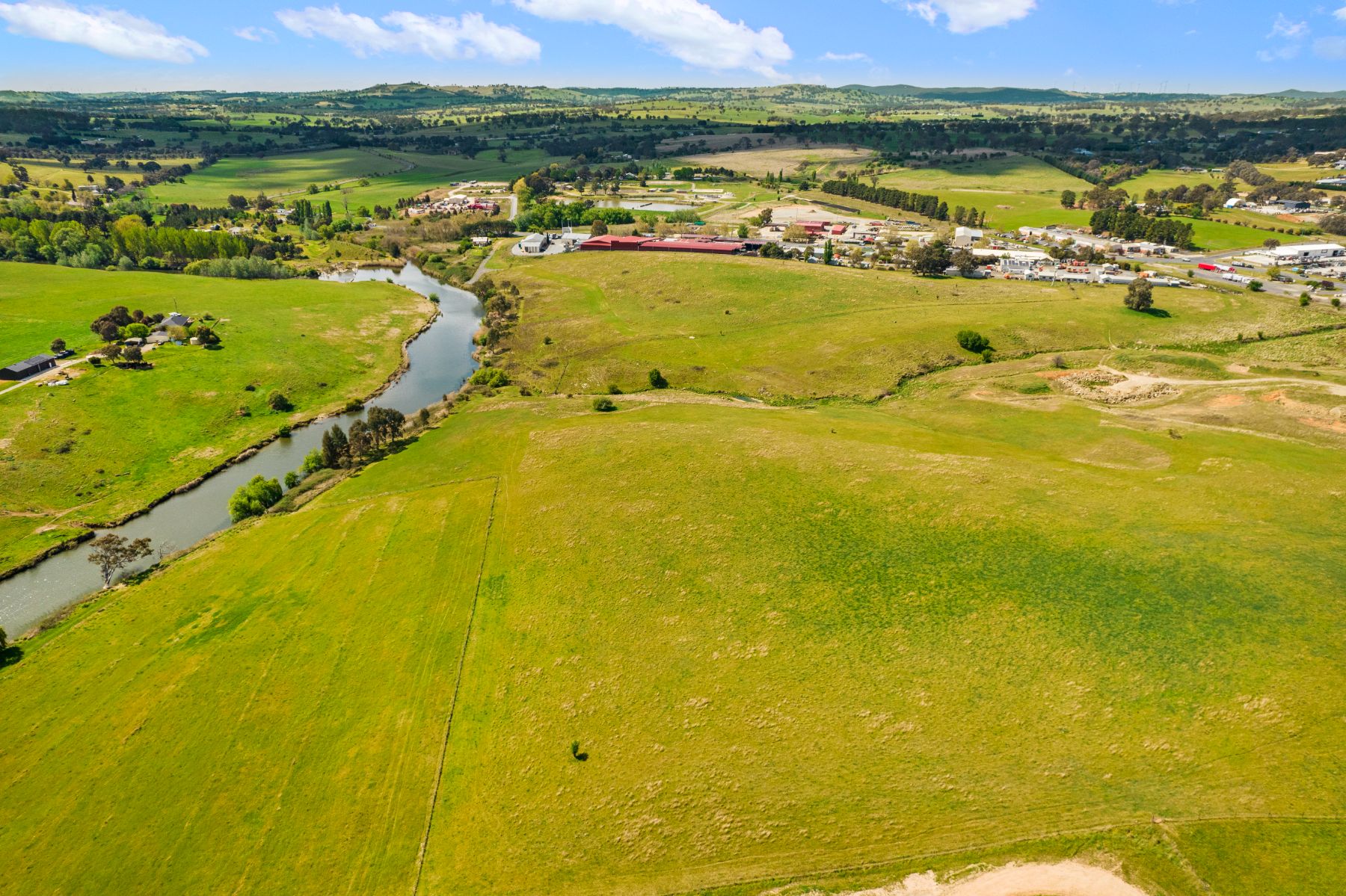 Aerial view of open green paddocks running down to a narrow river lined with trees, with farm buildings and a commercial area in the distance.