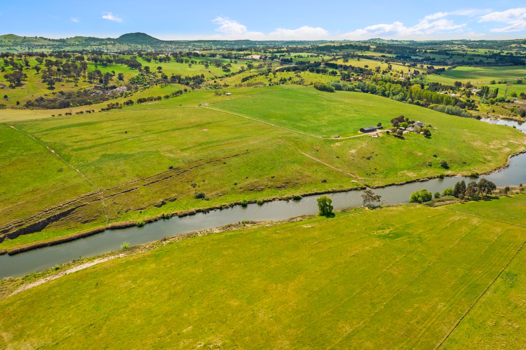 Aerial view of green paddocks on a bend of a river, with a farmhouse and sheds on the far bank and wooded hills behind.